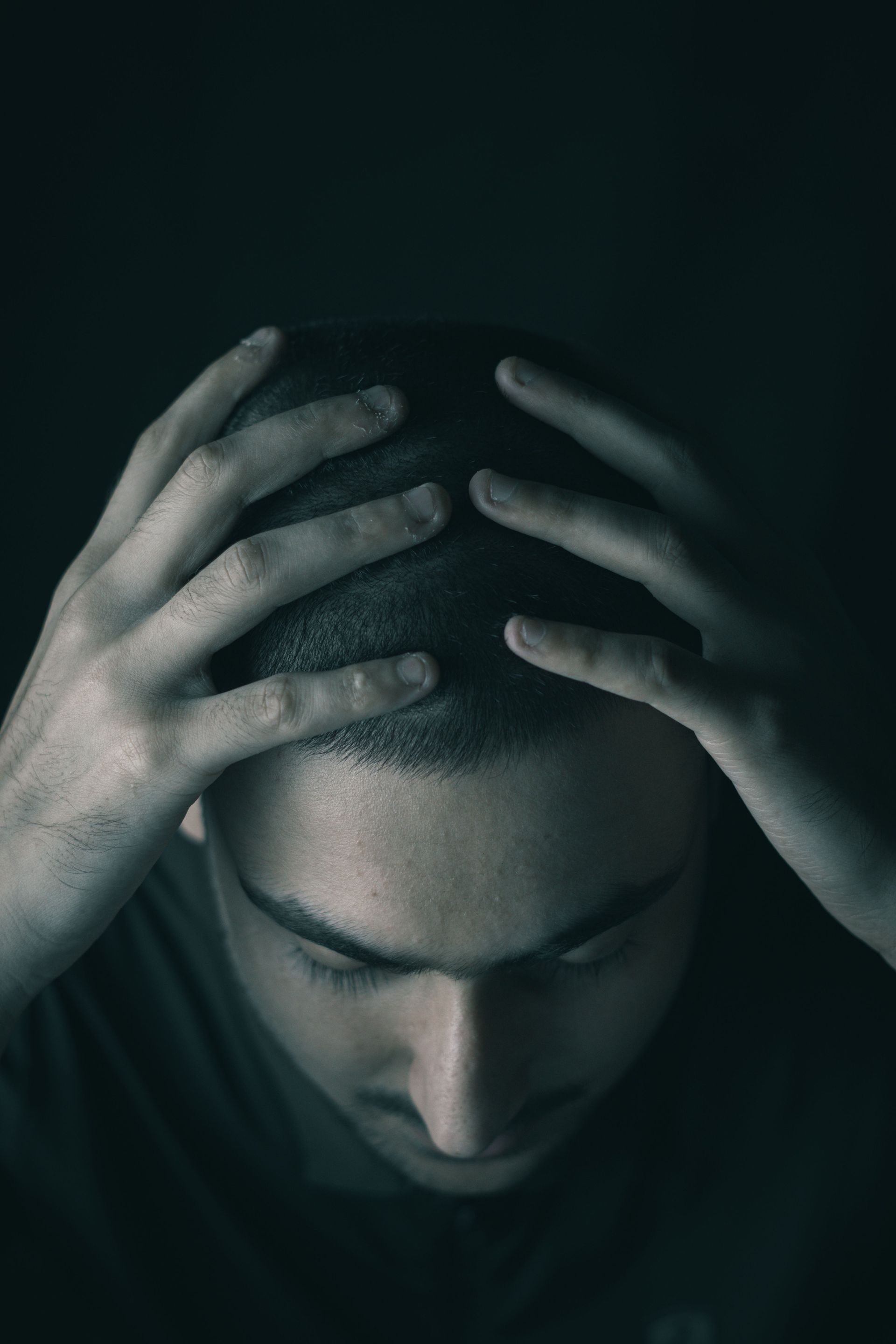 A man sitting in a dark room, hands on head, conveying stress and depression.