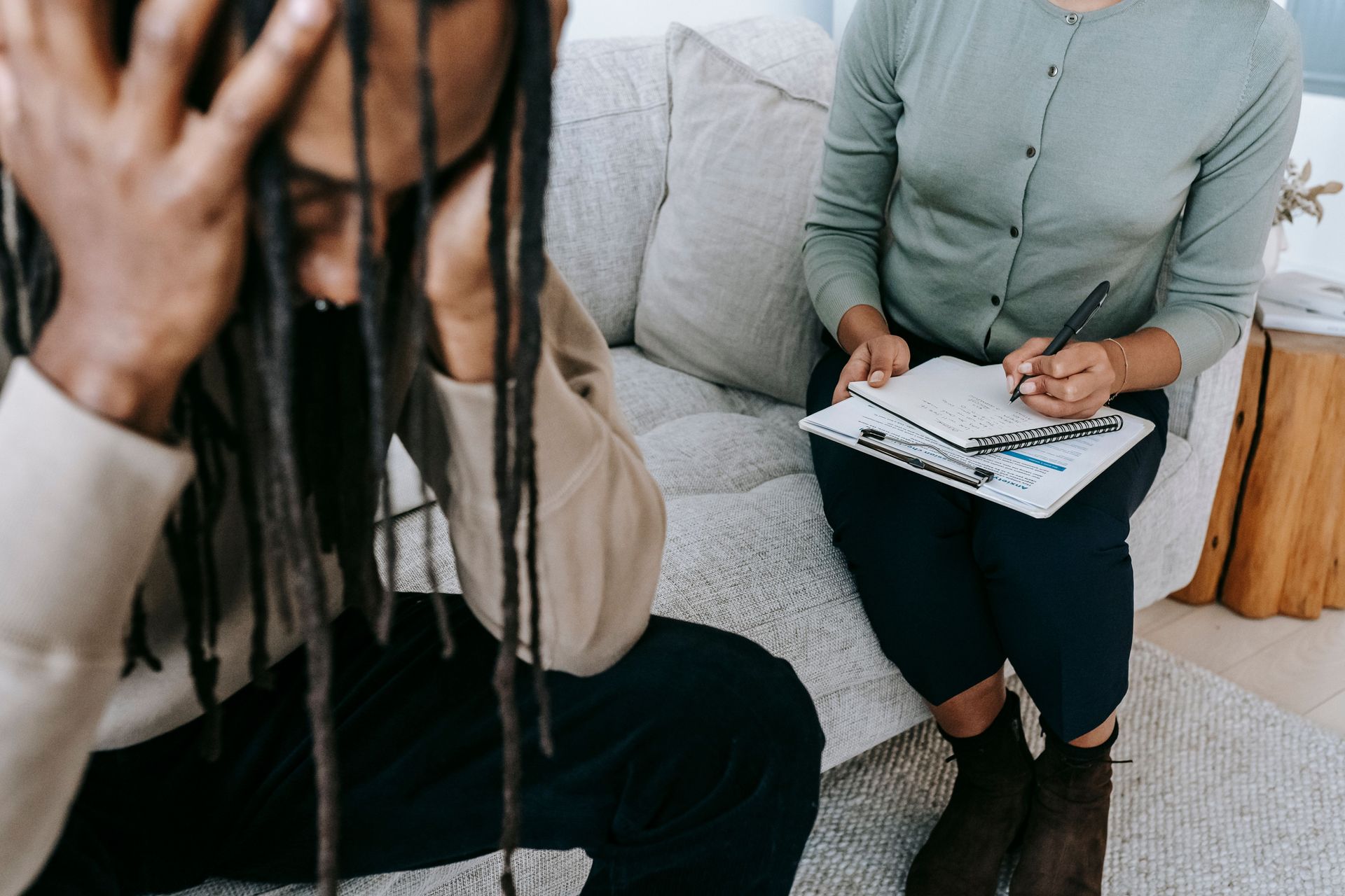 Crop concerned African American male sitting on sofa with female psychologist and holding head in hands in dismay during psychotherapy session