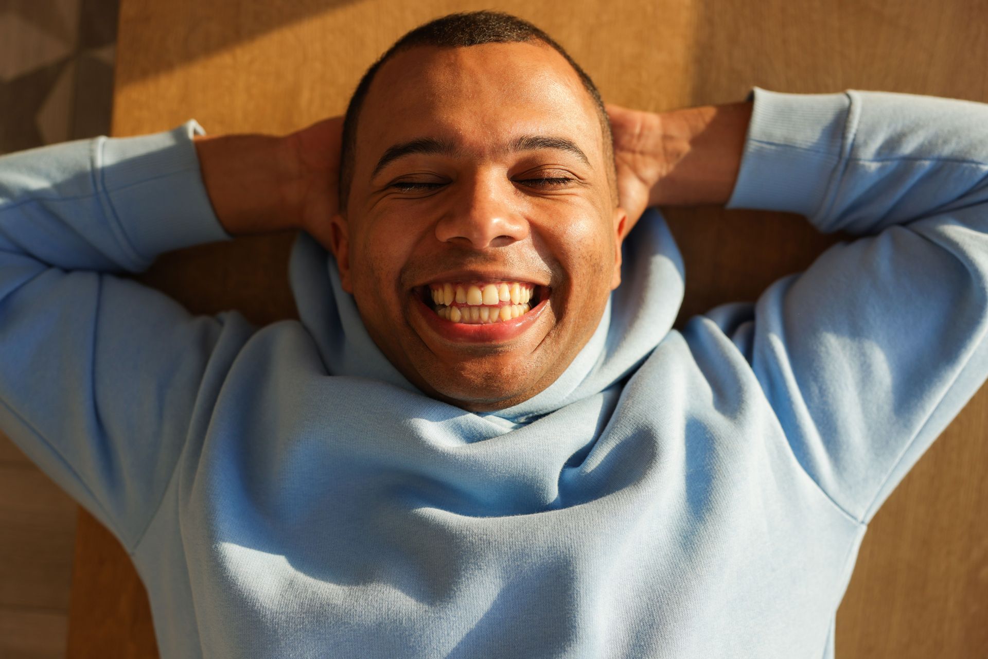 Close-up of a cheerful man in a blue hoodie lying down, smiling with eyes closed.