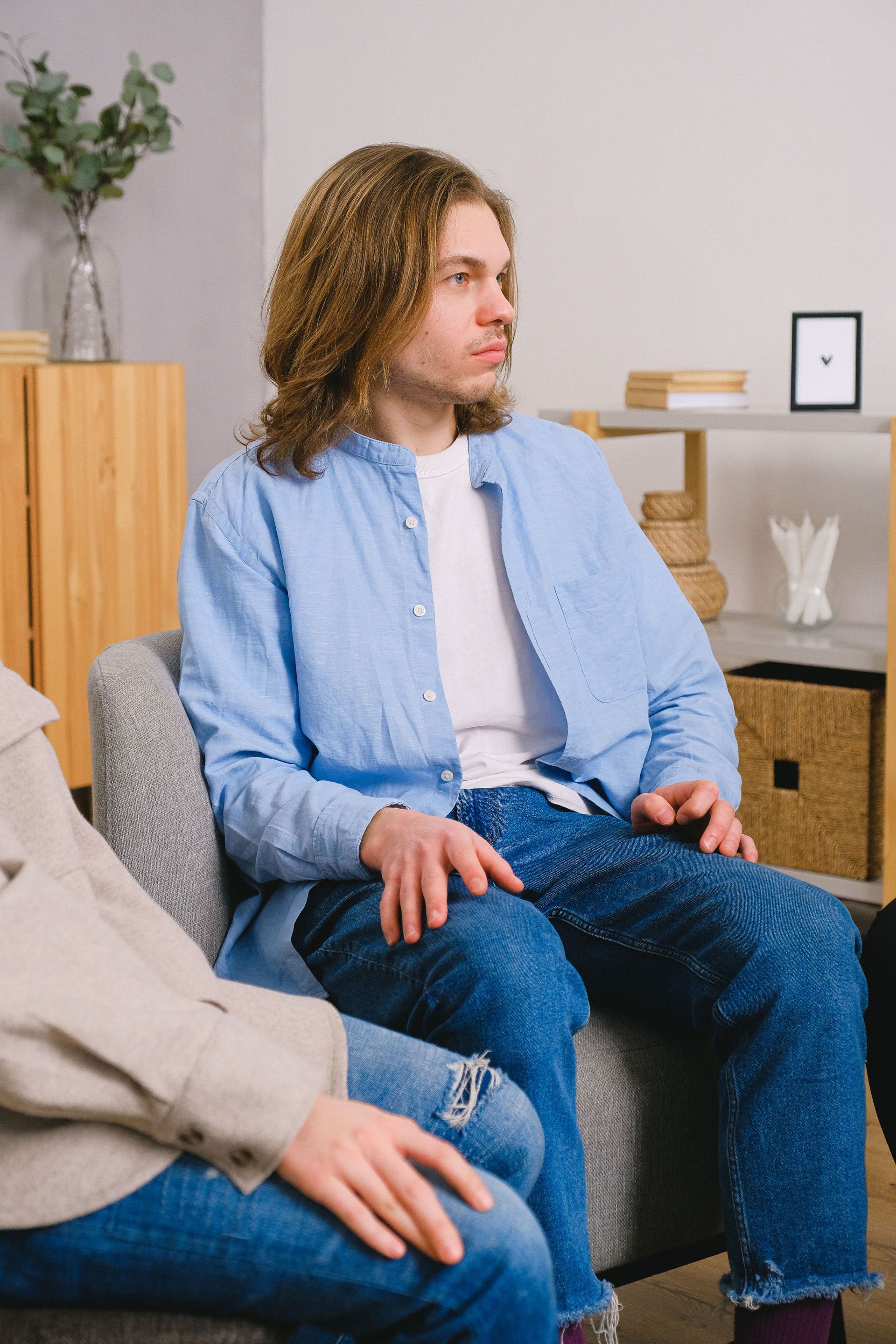 Long haired man in in shirt and jeans sitting on chair and looking away near unrecognizable partner during therapy session